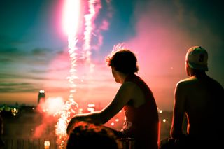 Two people in right foreground watch a stream of fireworks launch in the left background