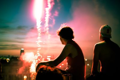 Two people in right foreground watch a stream of fireworks launch in the left background