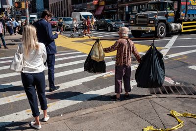 Woman on city street corner balancing several large garbage bags attached to a pole across her shoulders